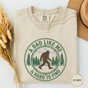 May include: Ivory-colored t-shirt with a Bigfoot graphic and the text "A DAD LIKE ME IS HARD TO FIND" in a green circular design. The shirt features a crew neck and the Comfort Colors brand label.