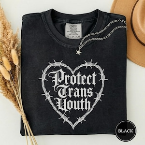 Puede incluir: Camiseta negra con un diseño blanco en forma de corazón hecho de alambre de púas. Las palabras "Protect Trans Youth" están impresas en una fuente gótica dentro del corazón. Un collar plateado con un colgante de estrella descansa sobre la camiseta.