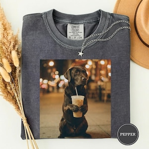 Könnte beinhalten: Dunkelgraues Comfort Colors T-Shirt mit einem Foto eines braunen Hundes, der einen Eiskaffee hält. Der Hund sitzt auf einem Bürgersteig mit verschwommenem Hintergrund einer Straßenszene. Eine silberne Sternkette ist auf dem Shirt drapiert.