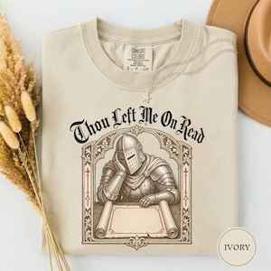 May include: An ivory t-shirt featuring a graphic of a knight in armor resting their head on one hand while leaning over a scroll. The text above the illustration reads Thou Left Me On Read in a black gothic font. The shirt has a crew neckline.