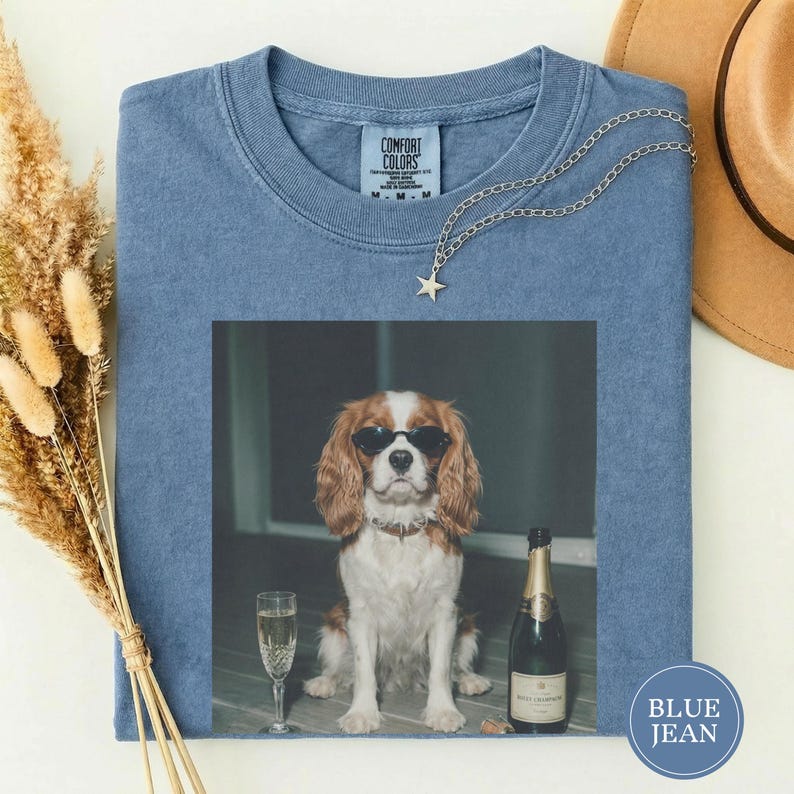 May include: A blue short-sleeved t-shirt with a Comfort Colors label at the neck. The center features a graphic of a brown and white spaniel wearing black sunglasses, sitting next to a champagne flute and a bottle of champagne.