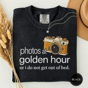May include: Black t-shirt with the text "photos at golden hour or i do not get out of bed." The shirt features a graphic of a vintage camera in tan and gold. A silver necklace with a star pendant is draped on the shirt.