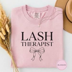 以下が含まれることがあります： 「LASH THERAPIST」の文字とリボンがプリントされた、薄いピンク色のComfort ColorsのTシャツ。黒い文字で書かれています。星のペンダントが付いたシルバーのネックレスと茶色の帽子が見えます。Tシャツは折りたたまれています。