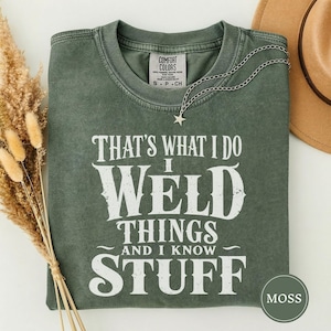 May include: A moss green crewneck t-shirt with a distressed white graphic print that reads, "THAT'S WHAT I DO I WELD THINGS AND I KNOW STUFF." The shirt features a Comfort Colors brand label at the inner neckline and a silver star-shaped necklace.