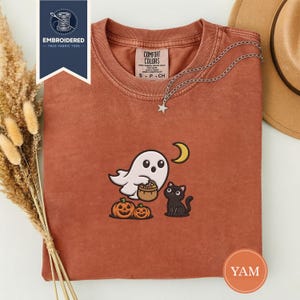Puede incluir: Camiseta de color óxido con un diseño de Halloween bordado. El diseño presenta un fantasma blanco sosteniendo una cesta, un gato negro, dos calabazas y una luna creciente. Un collar plateado con un colgante de estrella está sobre la camiseta.