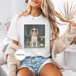 May include: A white t-shirt featuring a graphic print of a brown and white spaniel dog wearing black sunglasses, sitting next to a bottle of champagne and a filled champagne flute. The shirt has a round neckline and short sleeves.