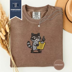 May include: Espresso-colored t-shirt with an embroidered raccoon holding a yellow clipboard and pointing upwards. The shirt has a crew neck and a Comfort Colors tag. A silver necklace with a star pendant is draped on the shirt. The image also includes a brown hat and dried plants.