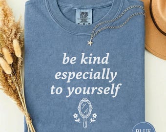 Könnte beinhalten: Blaues Jeans-T-Shirt mit dem weißen Text "be kind especially to yourself". Eine silberne Halskette mit Sternanhänger liegt auf dem Shirt. Ein brauner Hut ist im Hintergrund.