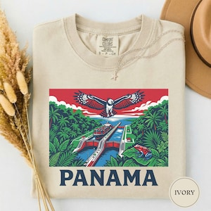 May include: Ivory-colored t-shirt featuring a vibrant graphic of the Panama Canal with an eagle, lush greenery, and a red frog. The word "PANAMA" is printed in bold blue letters below the image. The shirt is a Comfort Colors brand.