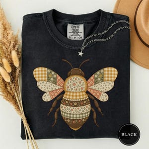 以下が含まれることがあります： 黒のクルーネックTシャツ。蜂の大きなグラフィックが描かれており、その羽と体は、花柄、ギンガムチェック、水玉模様など、茶色、クリーム色、くすんだ緑色の様々なパッチワークパターンで構成されています。