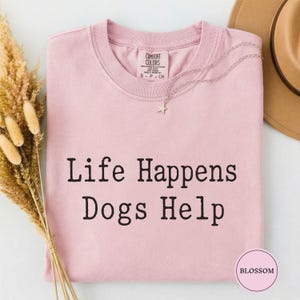 May include: A light pink short-sleeved t-shirt with the black text "Life Happens Dogs Help" printed on the front. The shirt features a crew neckline and a Comfort Colors brand label on the inside collar. A silver star pendant necklace rests on the fabric.
