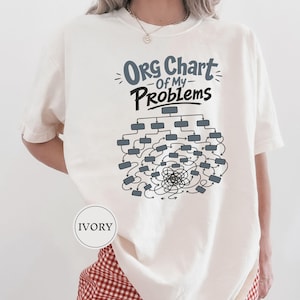 May include: An ivory short-sleeve t-shirt featuring a graphic print with the text "ORG Chart Of My Problems" in blue and black. Below the text is a complex organizational chart diagram with numerous interconnected boxes and a tangled center.