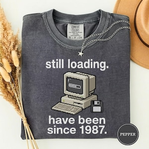 May include: A dark gray t-shirt with the text "still loading." and "have been since 1987." A pixelated computer graphic is in the center. A silver necklace with a star pendant is draped on the shirt.
