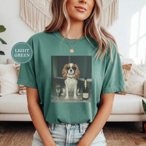May include: A light green short-sleeve t-shirt featuring a graphic print of a brown and white spaniel dog wearing black sunglasses, sitting next to a champagne flute and a bottle of champagne. The text LIGHT GREEN is printed in a circular badge.