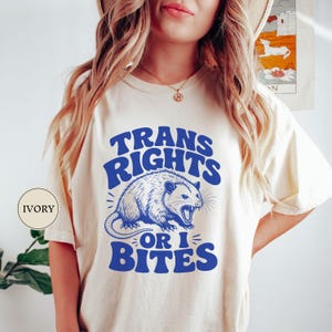 May include: Ivory-colored t-shirt with the text "TRANS RIGHTS OR I BITES" in blue, along with a blue illustration of an opossum. The shirt is a casual wear item.