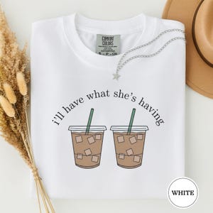 May include: White t-shirt with the phrase "I'll have what she's having" and two iced coffee illustrations. The shirt has a crew neck and a tag that says "Comfort Colors". A silver necklace with a star pendant is draped on the shirt.