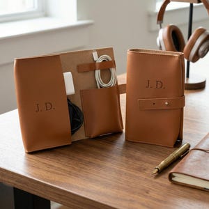 May include: A collection of brown leather tech organisers and a matching notebook on a wooden surface. The organisers are embossed with the initials "J.D." and include a cable organiser and a snap-closure case. A pen and headphones are also visible.