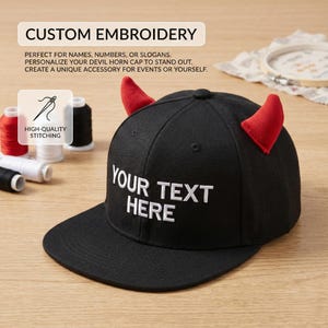 May include: Black baseball cap with red devil horns and the text "YOUR TEXT HERE" embroidered in white. The cap features a flat brim and a structured crown. The image also shows spools of thread and the words "CUSTOM EMBROIDERY".