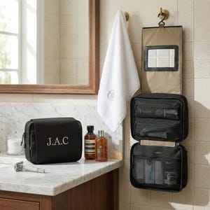 Könnte beinhalten: Eine schwarze Kulturtasche mit den Initialen "J.A.C" in Weiß gestickt. Die Tasche steht auf einer Marmortheke neben zwei braunen Flaschen und einem silbernen Rasierer. Ein weißes Handtuch hängt an einem Messinghaken.