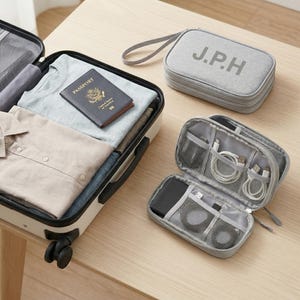 Könnte beinhalten: Ein offener Koffer mit Kleidung und einem Reisepass auf einer Holzoberfläche. Ein grauer Reiseorganizer mit den Initialen "J.P.H" neben einem offenen grauen Technik-Organizer mit Kabeln und Zubehör. Reise- und Organisationskonzept.