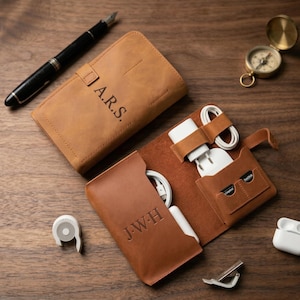 May include: A brown leather tech organizer with the initials "A.R.S." and "J.W.H." engraved on the exterior. The organizer is open, revealing compartments for chargers, cables, and SD cards. A black pen and a compass are also visible.