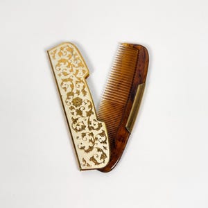May include: An open, antique comb with a decorative ivory-coloured handle featuring gold filigree. The comb itself is a dark amber colour with fine teeth. A small brass hinge connects the handle and comb.