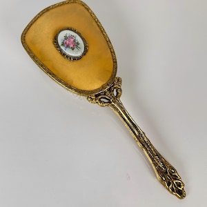 Könnte beinhalten: Eine goldfarbene Haarbürste mit einem verzierten, strukturierten Griff und einer ovalen Einlage in der Mitte, die ein Blumenmuster aus rosa Rosen und blauen Akzenten auf weißem Grund zeigt. Der Bürstenkopf hat eine glatte, bernsteinfarbene Oberfläche.