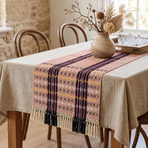 Peut inclure: Un chemin de table rectangulaire avec une base rose poudré, des bandes verticales violet foncé, des lignes en zigzag jaunes et des motifs géométriques blancs en forme de losanges et de croix. Les extrémités présentent de longues franges.