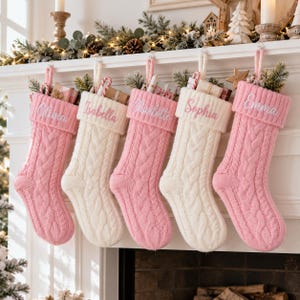 May include: Five cable-knit Christmas stockings in alternating pink and cream colours, each featuring a folded cuff embroidered with the names Olivia, Isabella, Charlotte, Sophia, and Emma. Each stocking contains small gift boxes and greenery.