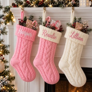May include: Three cable-knit Christmas stockings hanging from a mantel. Two are pink with white cuffs embroidered with the names Harper and Rosalie, and one is white with a white cuff embroidered with the name Lillian. Each contains greenery and gifts.