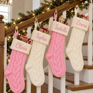 May include: Four cable-knit Christmas stockings hanging in a row. Two are pink with white cuffs and two are white with pink cuffs. Each cuff is embroidered with a name: Olivia, Sophia, Isabella, and Emma. Small gifts and candy canes are inside.