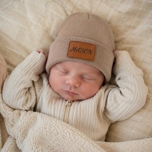 May include: A light beige knit beanie featuring a rectangular brown leather patch on the front. The patch is embossed with the name MASON in a dark, elegant script font. The beanie has a ribbed texture and a folded cuff design.