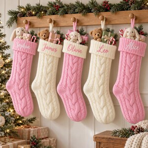 Op de afbeelding: Vijf kerstsokken van kabelgebreid materiaal hangen aan een houten rek. Drie zijn roze met witte boorden en twee zijn crème met crème boorden. Elke sok bevat een knuffel, een klein cadeautje en de geborduurde namen Sophia, James, Olivia, Leo en Alice.