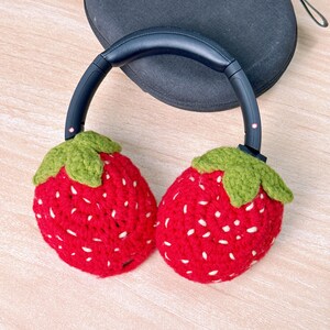 May include: Headphones with red, crocheted strawberry ear covers. The strawberries have white seed details and green leafy tops. The headphones have a black headband and earcups, and are resting on a black case.