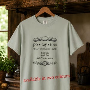 May include: A light sage green t-shirt featuring a black graphic of five potatoes above the text "po • tay • toes", "a field guide", and the instructions "boil 'em", "mash 'em", "stick 'em in a stew" with a small barrel illustration and leafy accents.