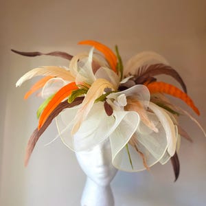 May include: A decorative headpiece with a white base, adorned with orange, brown, and cream-colored feathers. The headpiece also features white and cream-colored fabric accents. The design is displayed on a white mannequin head.