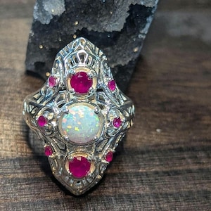 May include: Ornate silver ring featuring a central oval opal, flanked by two round rubies and accented with smaller rubies. The ring has an intricate filigree design, showcasing a vintage or antique style. The gemstones are set in a detailed, decorative setting.