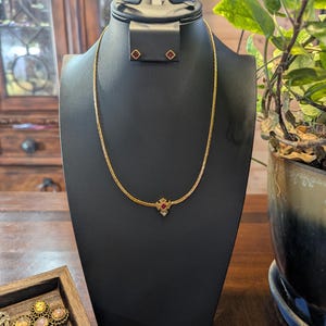 May include: A gold-toned necklace and matching earrings set displayed on a black jewelry bust. The necklace features a small, red gemstone pendant. The earrings are small, square-shaped, and also feature red gemstones. The jewelry is presented on a wooden surface.