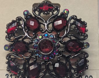 May include: A vintage brooch with a dark metal filigree design, featuring oval and round red gemstones. The brooch has iridescent accents and is set on a cream-colored card with the text "1928."