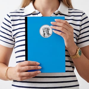 Puede incluir: Un cuaderno de color azul brillante con un lomo negro y un emblema circular centrado. El emblema contiene un dibujo lineal detallado en blanco y negro de una persona con un tocado de plumas, presentado en un estilo ilustrativo clásico.