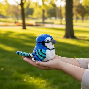 Pode incluir: Um brinquedo de pelúcia de gaio-azul feito à mão, com barriga branca, detalhes em preto e bico cinza. O pássaro é feito de crochê com um padrão detalhado e é segurado em mãos abertas.