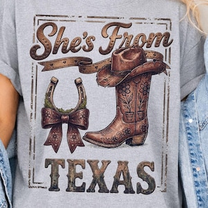 May include: A gray t-shirt with a graphic that reads "She's From Texas." The graphic includes a cowboy hat, a horseshoe with a bow, and a cowboy boot. The words "Instant Download" are at the bottom.