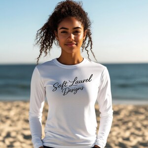 May include: A white long-sleeve shirt with the text "Soft Laurel Designs" in black script. The shirt is made of a lightweight material and is suitable for outdoor activities. The model is standing on a beach.