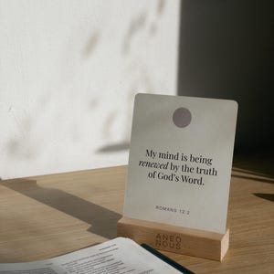 Könnte beinhalten: Eine Karte mit dem Text "My mind is being renewed by the truth of God's Word. Romans 12:2" wird in einem Holzhalter angezeigt. Die Karte hat einen hellvioletten Kreis oben. Ein offenes Buch befindet sich im Vordergrund.