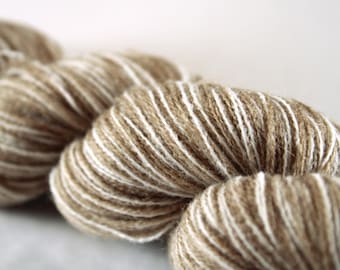 Op de afbeelding: Een streng garen met een getwiste constructie van een mix van lichtbruine, beige en helderwitte vezels. Het garen heeft een pluizig, getextureerd uiterlijk met zichtbare draden in contrasterende kleuren die samen in een lus zijn gewonden.