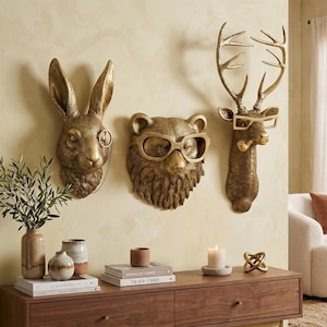 May include: Three gold-colored animal head wall decorations: a rabbit with a monocle, a bear with glasses, and a deer with glasses and a pipe. A wooden console table below holds books, vases, a candle, and a decorative knot.