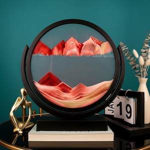 Könnte beinhalten: Ein rundes Sandkunstwerk in einem schwarzen Rahmen, das eine Berglandschaft in Rot- und Beigetönen darstellt. Die Kunst wird auf einem schwarzen Tisch zusammen mit einer goldenen Statue und einem Kalender ausgestellt.