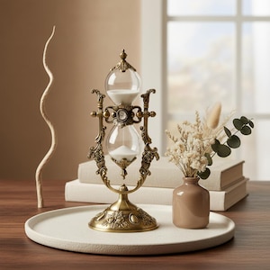 May include: An ornate hourglass with a brass finish. The hourglass features two glass bulbs connected by a narrow neck, filled with white sand. The decorative stand has floral and scrollwork details. A small vase with dried flowers sits nearby.