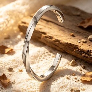 May include: A silver-colored, open-ended cuff bracelet with a twisted design. The bracelet is displayed on a white, textured surface with a wooden plank in the background. The bracelet's polished surface reflects light, creating highlights and shadows.