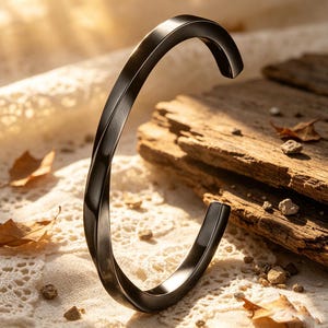 Könnte beinhalten: Ein schwarzes, verdrehtes Metallarmband. Das Armband hat eine glatte, polierte Oberfläche und ein geschwungenes Design. Der Hintergrund zeigt eine strukturierte weiße Oberfläche, Holzbretter und verstreute Blätter, die eine natürliche Ästhetik schaffen.